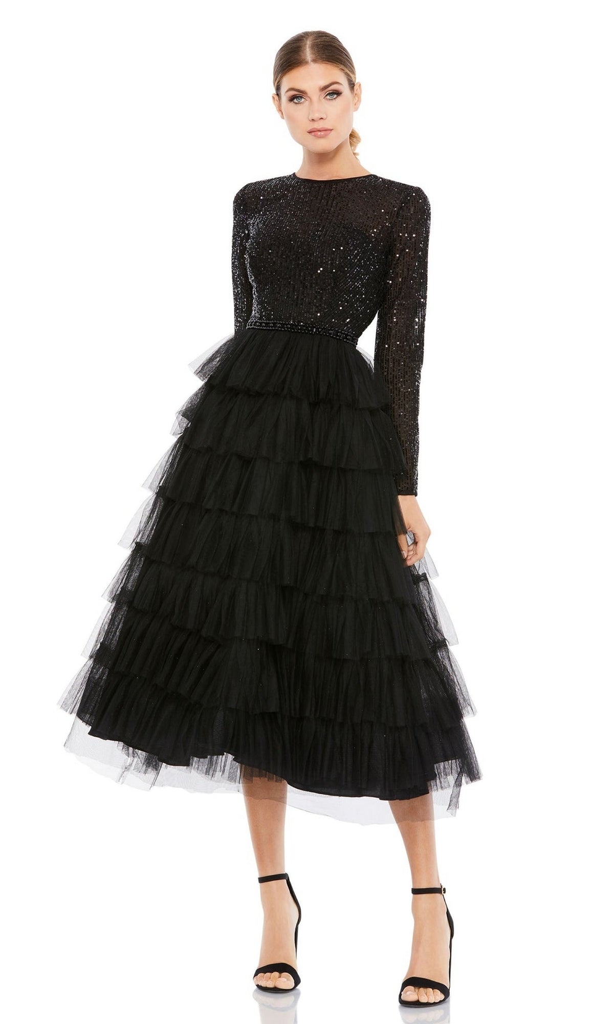 A woman wears the Mac Duggal 26299 knee-length wedding guest dress—a black sequin A-line design with long sleeves and a layered tulle skirt—paired with black high-heeled sandals, set against a white background.